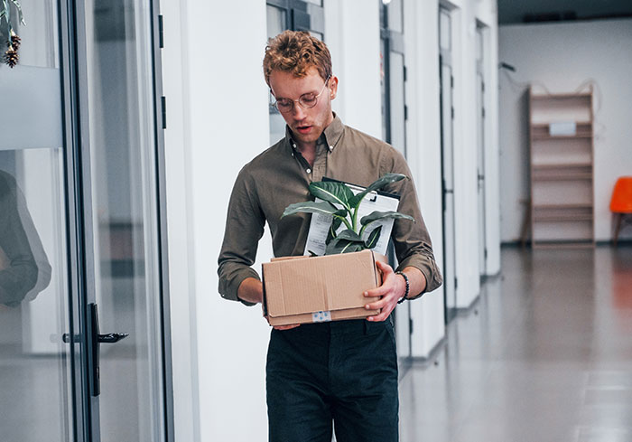 Employee carrying a box with a plant after being fired, showing workplace dynamics.