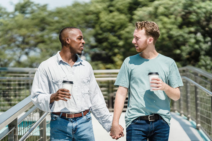 Two boyfriends holding hands and talking, each with a cup, walking outside; concept relates to mortgage discussion. Two boyfriends holding hands and talking, each with a cup, walking outside; concept relates to mortgage discussion.