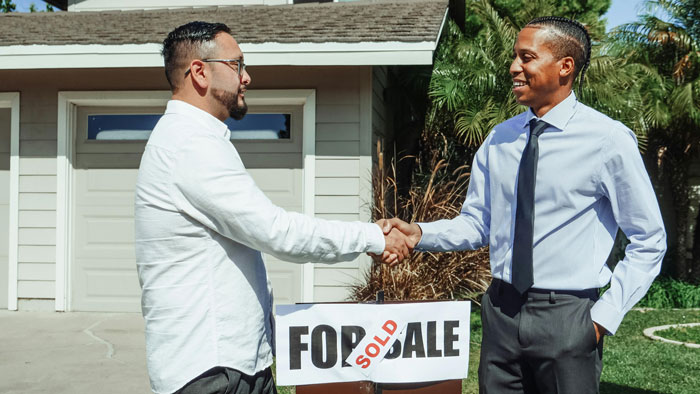 Two men shaking hands in front of a house with a "Sold" sign, discussing a mortgage agreement. Two men shaking hands in front of a house with a "Sold" sign, discussing a mortgage agreement.