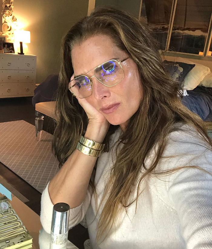 Brooke Shields wearing glasses sits indoors, resting her face on her hand, conveying contemplation.