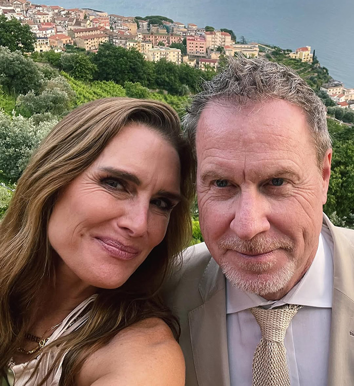 Brooke Shields with her husband smiling together in a scenic outdoor setting, overlooking a hillside town and the sea.