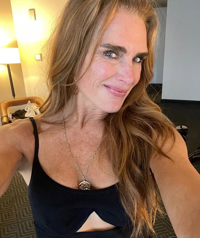 Brooke Shields with long hair and necklace, wearing a black top, smiling in a casual indoor setting.
