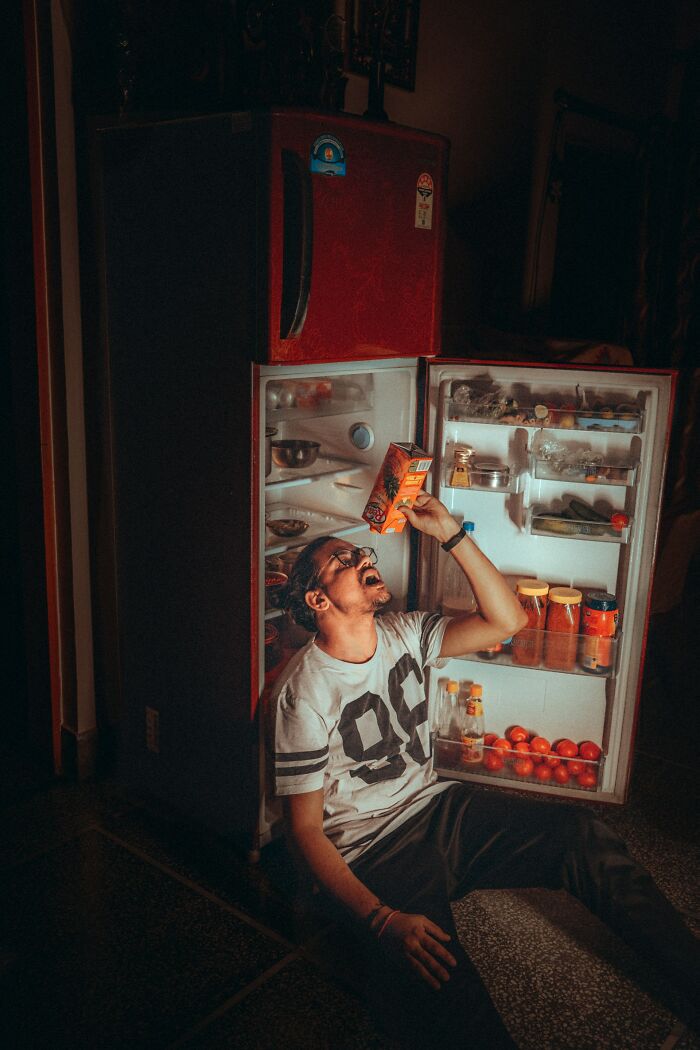 Man sitting by an open fridge, drinking juice directly from the carton, illustrating weight gain behavior.