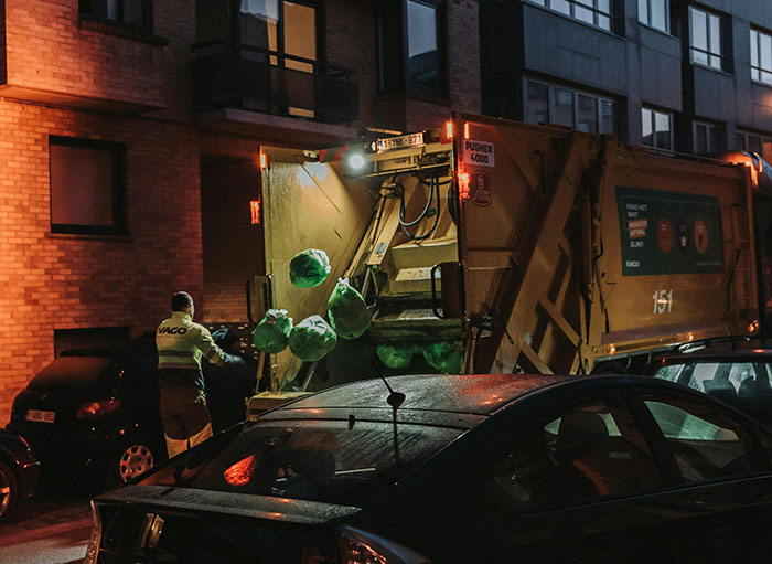Garbage truck at night with a collector loading bags, illustrating issues with unpaid overtime for waste workers. Garbage truck at night with a collector loading bags, illustrating issues with unpaid overtime for waste workers.