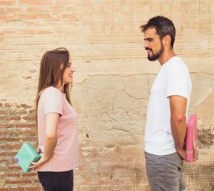 Woman and man smiling at each other outside, holding gifts behind their backs, discussing a neighbor interaction.
