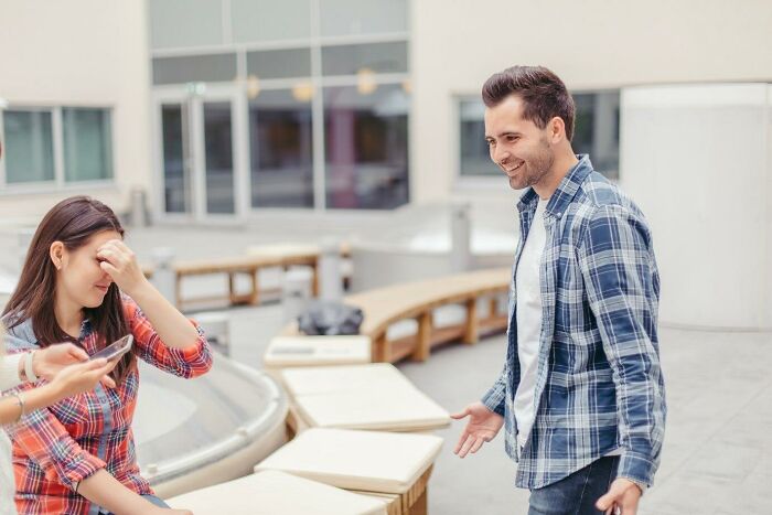Man smiling outdoors, wearing a plaid shirt, while a woman looks at her phone; neighbor interaction context.
