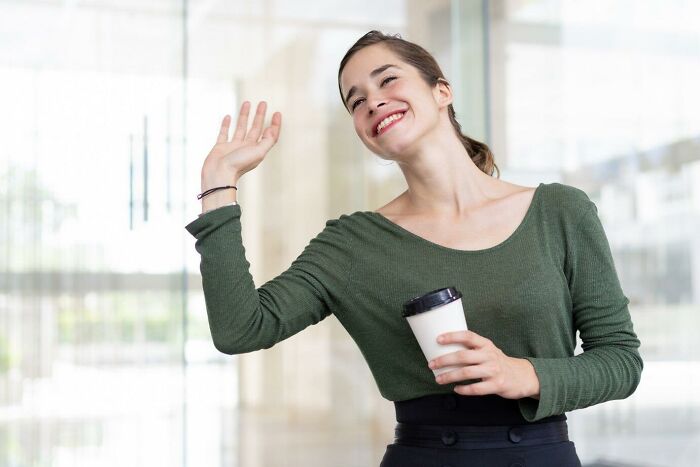 Woman in green sweater smiling and waving, holding a coffee cup; focusing on neighbor interaction.