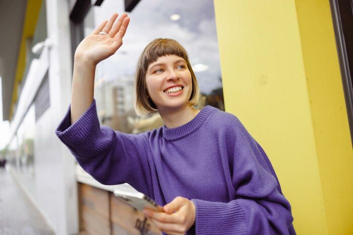 Woman smiling and waving while holding a smartphone, standing near a bright yellow wall.