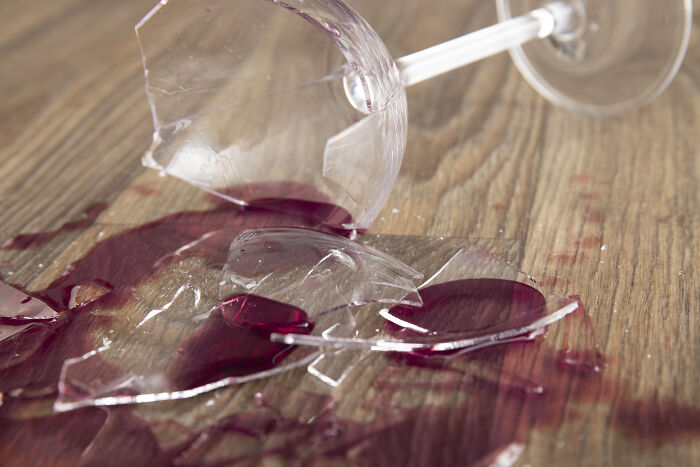 Broken wine glass with red wine spill on wooden floor.