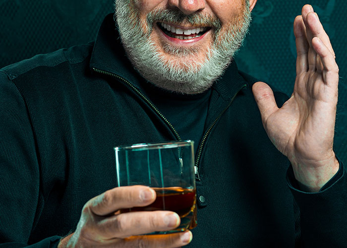 Older man with a gray beard smiling while holding a glass of whiskey, hinting at family secrets in a cozy setting.