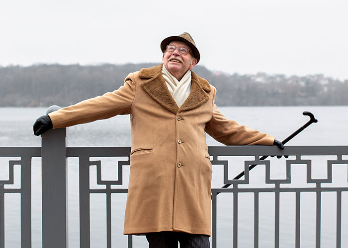 Elderly man in a brown coat and hat standing by a lake railing, smiling and holding a cane, reflecting on family secrets.