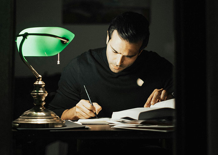 Man studying late at night under a green desk lamp, focused on uncovering horror movie-worthy family secrets.