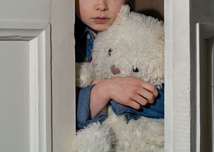 Child clutching a teddy bear, peeking from behind a door, evoking horror movie-worthy family secrets and hidden fears.