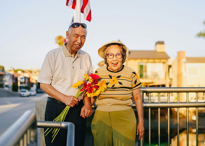 Elderly couple holding flowers outdoors, smiling and enjoying a sunny day, representing family secrets and relationships.