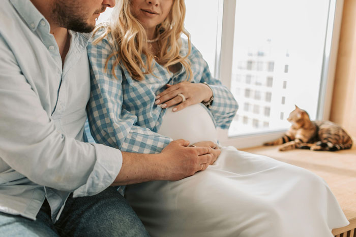 Dad and expectant mother sitting together, hands on her belly, with a cat by the window. Dad and expectant mother sitting together, hands on her belly, with a cat by the window.