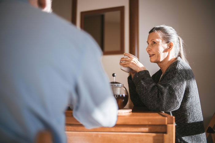 A woman in a sweater, seated at a table with a coffee pot, talking to a man, capturing a moment of connection. A woman in a sweater, seated at a table with a coffee pot, talking to a man, capturing a moment of connection.