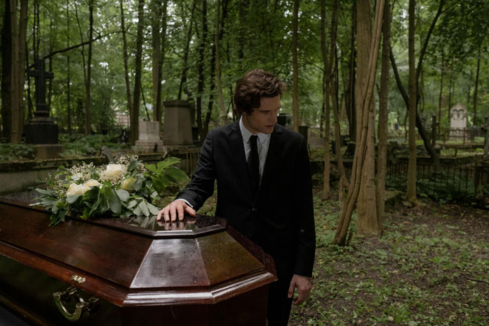 A man in a suit mournfully stands by a casket in a wooded cemetery, symbolizing dad grief. A man in a suit mournfully stands by a casket in a wooded cemetery, symbolizing dad grief.