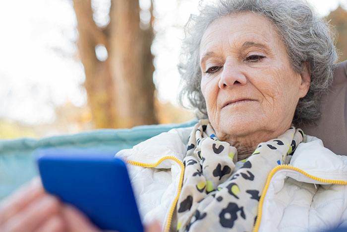 Elderly woman using smartphone outdoors, wearing scarf and jacket, reflecting on family and tuition promise. Elderly woman using smartphone outdoors, wearing scarf and jacket, reflecting on family and tuition promise.