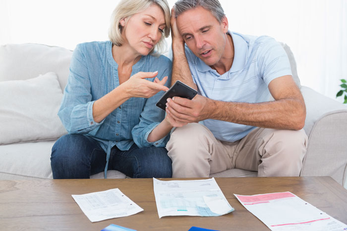 Parents looking stressed, sitting on a couch, surrounded by financial documents and reviewing a phone together.