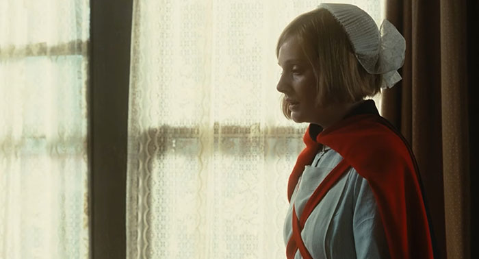 A woman in period clothing stands by a window, with light filtering through lace curtains, capturing a sad movie scene.