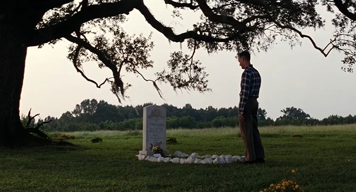 A man stands solemnly at a grave under a large tree, depicting a sad movie ending.