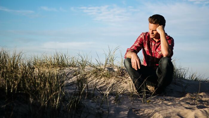 A man in a red plaid shirt sitting thoughtfully on a grassy sand dune, symbolizing intelligence signs like self-doubt.