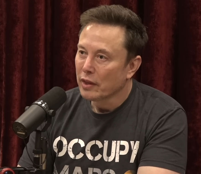 Man in "Occupy Mars" shirt, speaking into a microphone, against a red curtain backdrop. Man in "Occupy Mars" shirt, speaking into a microphone, against a red curtain backdrop.