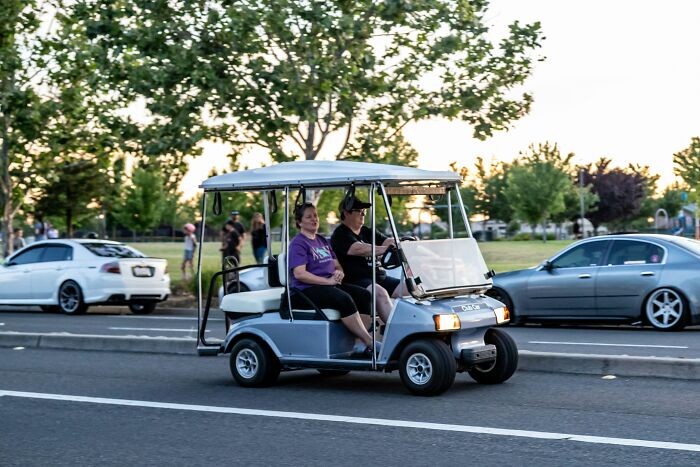 Two people in a golf cart driving on a street, choosing comfort over trends.