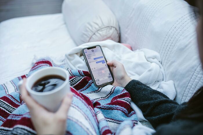 A woman holding a smartphone, texting, while sipping coffee on a cozy couch.