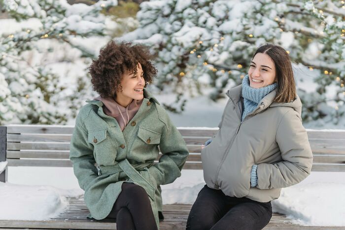 Two women in winter coats chatting on a snowy bench, symbolizing friendship and connection.