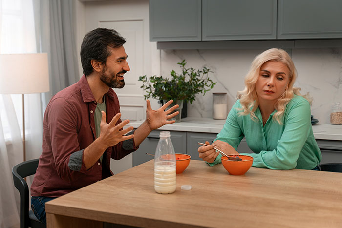 Husband speaking animatedly, wife looking away, sitting at a kitchen table with orange bowls and a milk bottle. Husband speaking animatedly, wife looking away, sitting at a kitchen table with orange bowls and a milk bottle.