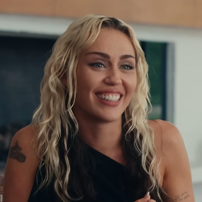 Miley Cyrus smiling with wavy blonde hair, wearing a one-shoulder top, in a casual indoor setting.