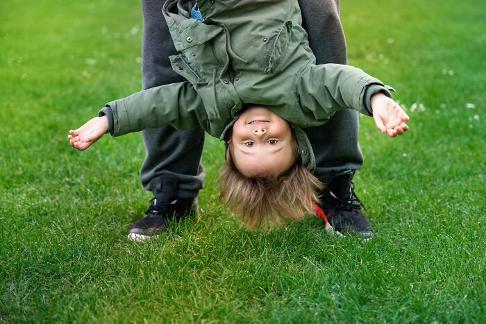Child in a green jacket smiling while upside down on grass, held by an adult in casual clothing.