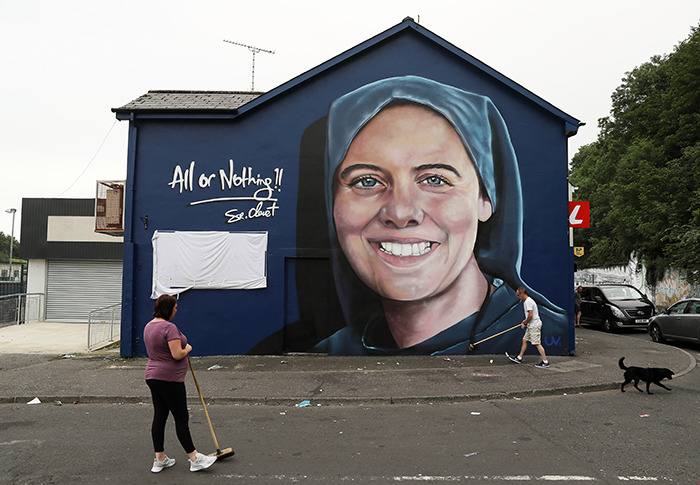 Mural of Sister Clare Crockett on a blue building, with people walking by. Mural of Sister Clare Crockett on a blue building, with people walking by.