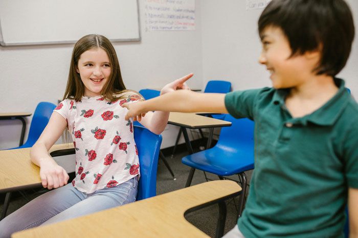 Two children pointing at each other, seated in a classroom with blue chairs, laughter in the air.