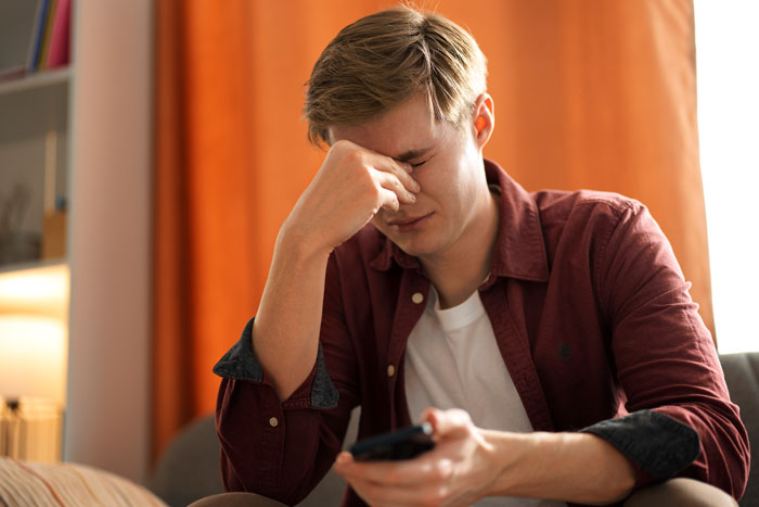 Teen holding phone, looking upset, wearing a maroon shirt, sitting in a room with orange curtains.