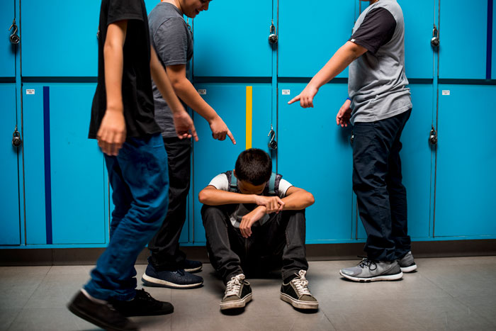 Teen enduring bullying, sitting by lockers, surrounded by peers pointing.