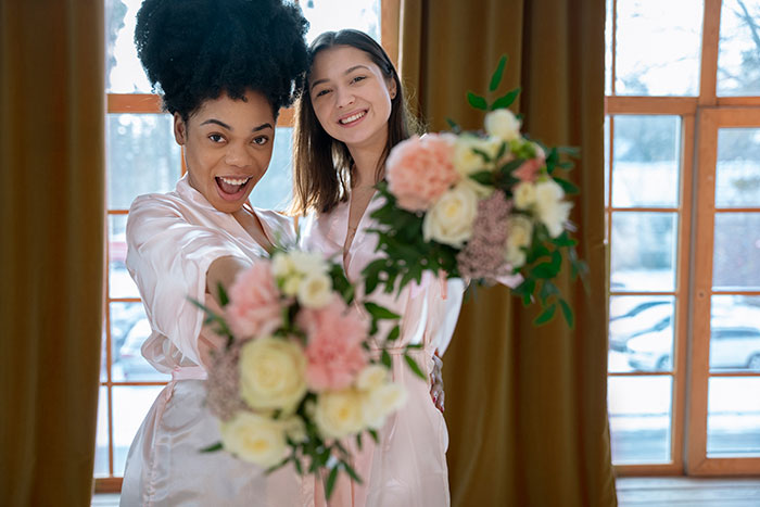 Two women in robes joyfully holding floral bouquets indoors near large windows.