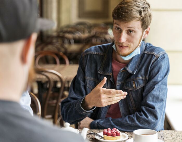 "Man in denim jacket gestures while discussing maturity over coffee and dessert at a café."