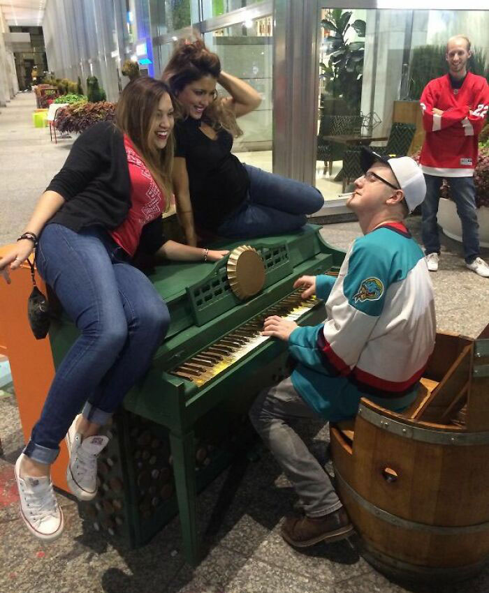 Man playing a green piano in a funny setting, with two women laughing near him, creating a humorous camera roll moment.