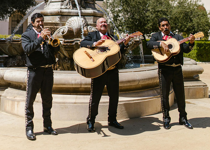 Musicians playing guitars and trumpet by a fountain outdoors, wearing traditional attire under sunlight.