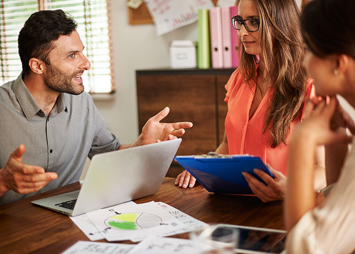 Man explaining a topic at a meeting while two women listen, one holding a clipboard, highlighting the concept of mansplaining.
