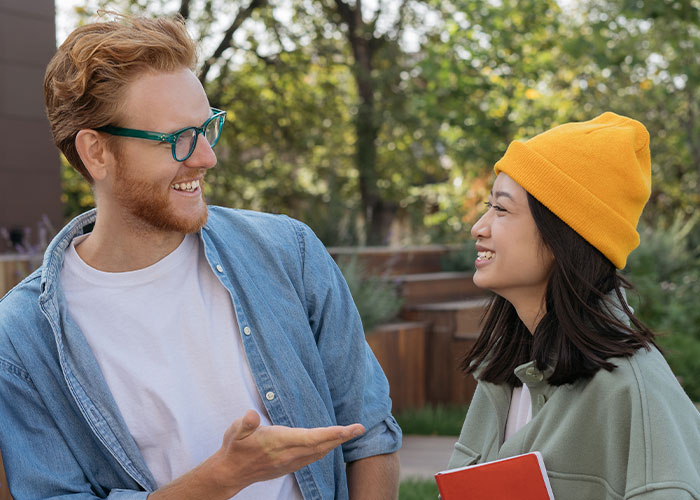 Man casually explaining something to a woman outdoors while she smiles, holding a red notebook.