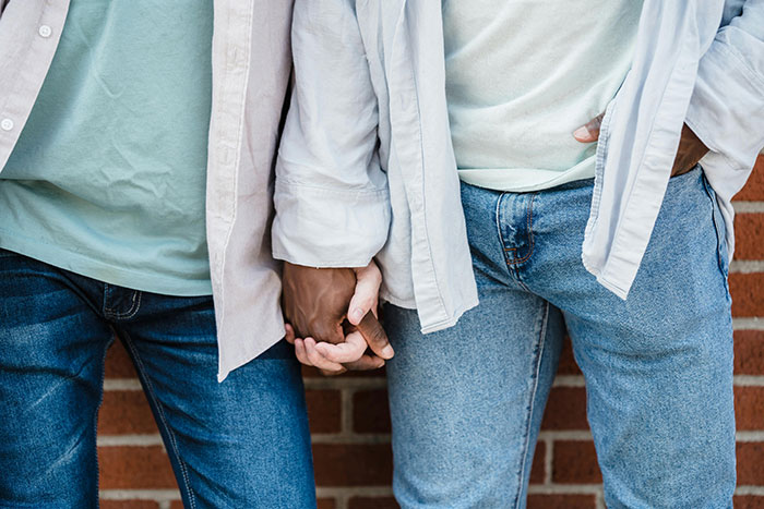 Two people in casual attire standing close, holding hands, with one wearing jeans and a white shirt, near a brick wall.