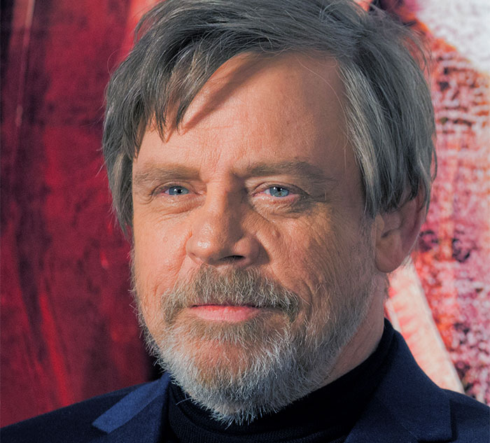 A celebrity with a beard and gray hair, standing against a red backdrop, looking thoughtful.