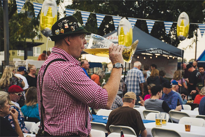 A man in a traditional Bavarian outfit drinking beer at a German festival, surrounded by people and festive decorations.
