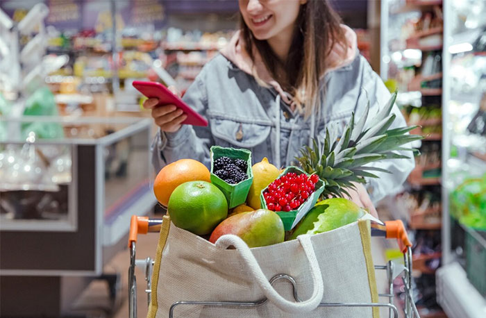 Woman in a grocery store with fruits in a cart, using a phone. Cultural insights from Germany influence her shopping habits.