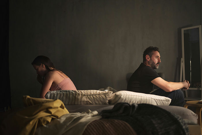 Man and woman sitting back to back on a bed, appearing upset, in a dimly lit room. Man and woman sitting back to back on a bed, appearing upset, in a dimly lit room.