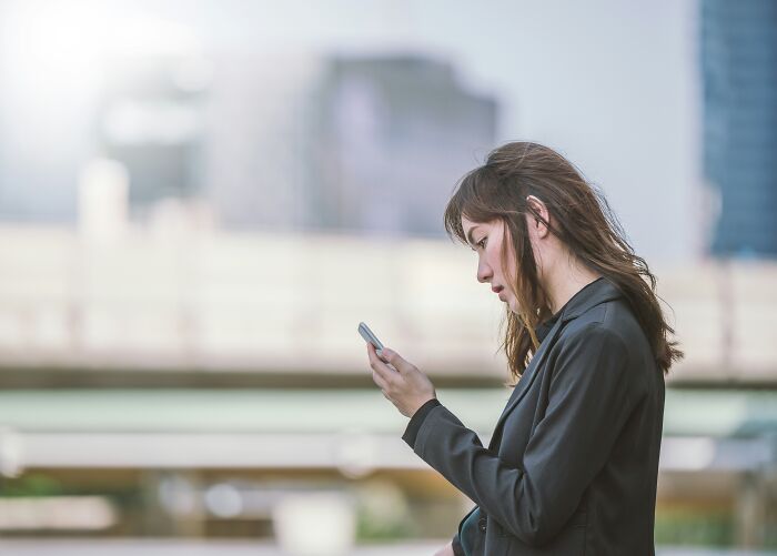 Woman in a black jacket looking at her phone, standing outdoors; signs of fake maturity.