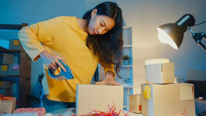 A woman in a yellow sweater uses a tape dispenser to seal a cardboard box, showcasing affordable life hacks.